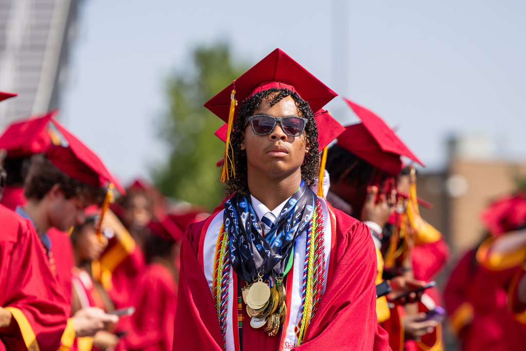 A Putnam City North graduate wearing medals, cords and sunglasses stands among classmates during graduation.