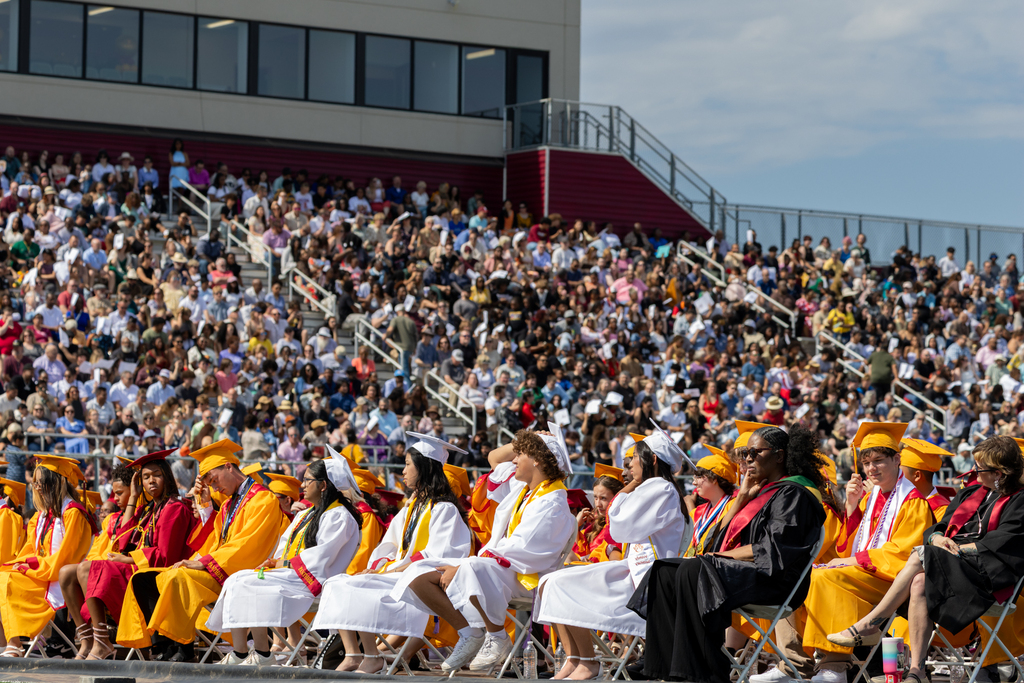 Putnam City North graduates sit in rows on the field with family and friends watching from the stadium bleachers.