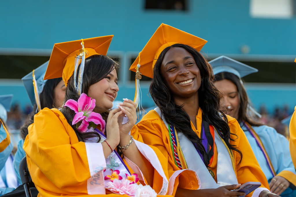 Two Putnam City West graduates in gold caps and gowns smile while seated with classmates during graduation.
