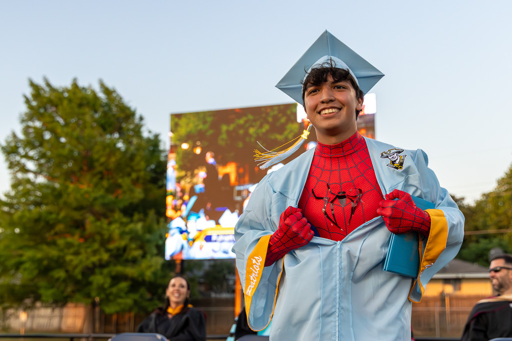 A Putnam City West graduate dressed as Spider-Man smiles while posing on stage in a cap and gown.