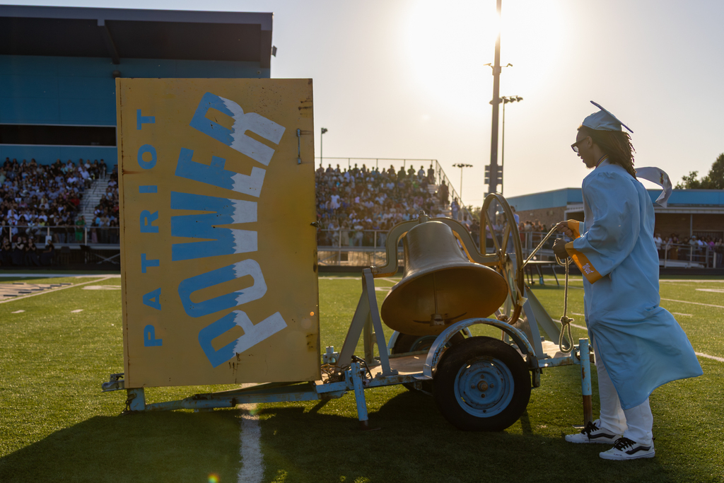 A Putnam City West graduate in a light blue cap and gown rings the Patriot Power bell on the field.