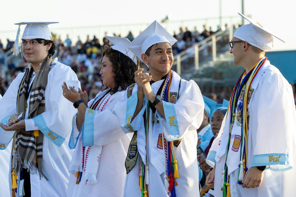 Putnam City West graduates in white caps and gowns applaud during the graduation ceremony.
