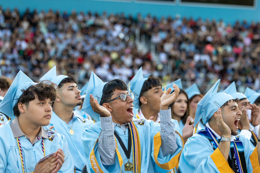 Putnam City West graduates in light blue caps and gowns cheer from their seats during the ceremony.