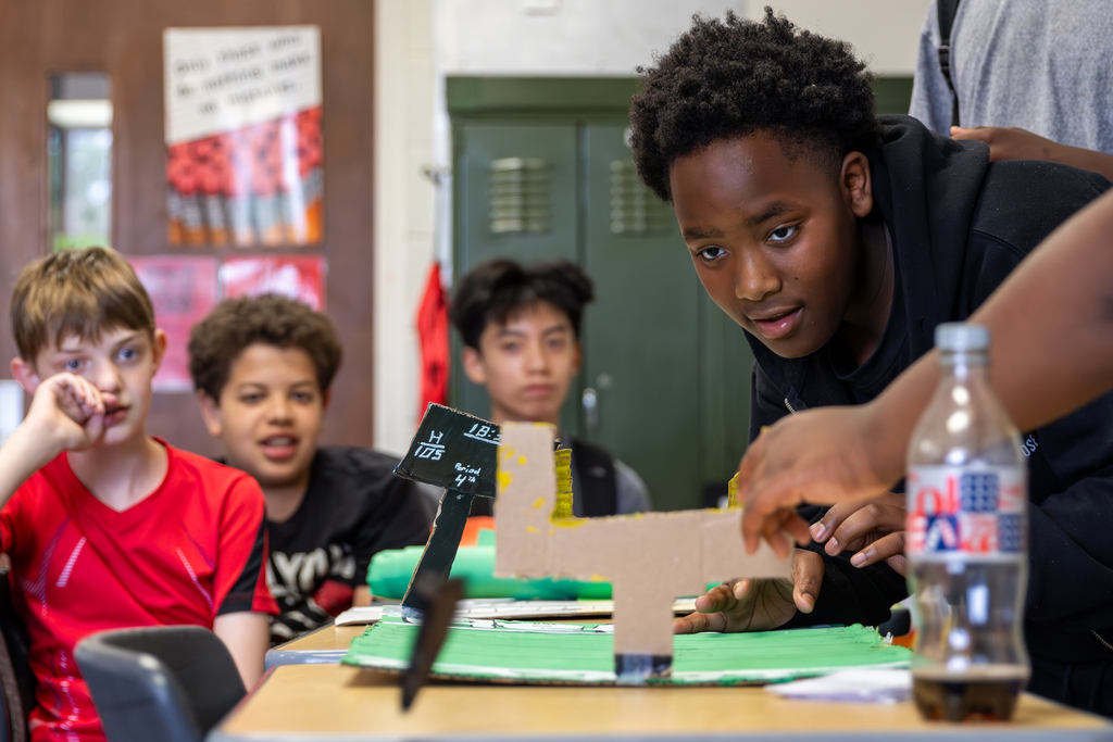 A Hefner Middle School student leans over a handmade board game while classmates watch during a math final project presentation.