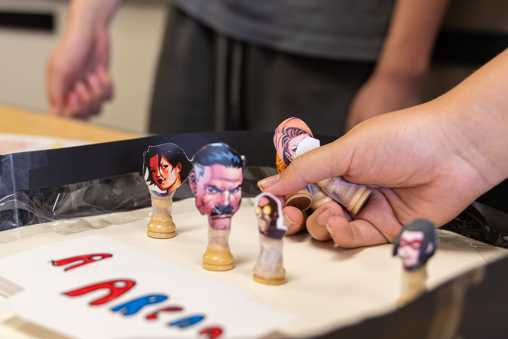 A close-up shows handmade character game pieces being moved on a student-created math board game.