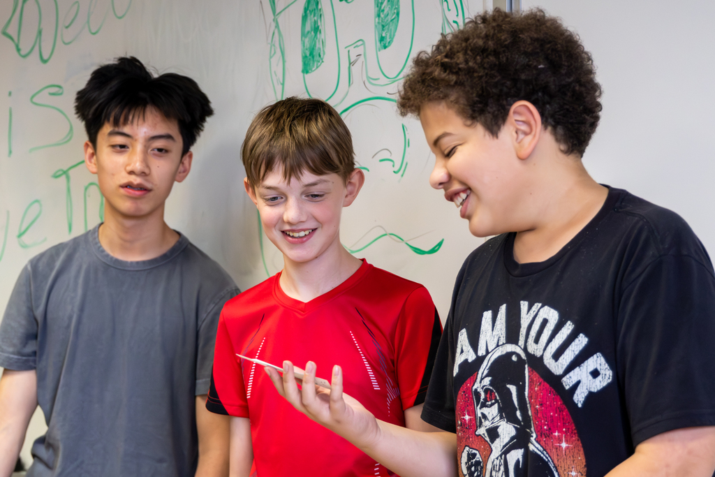 Three Hefner Middle School students smile while looking at game pieces during a math board game project.