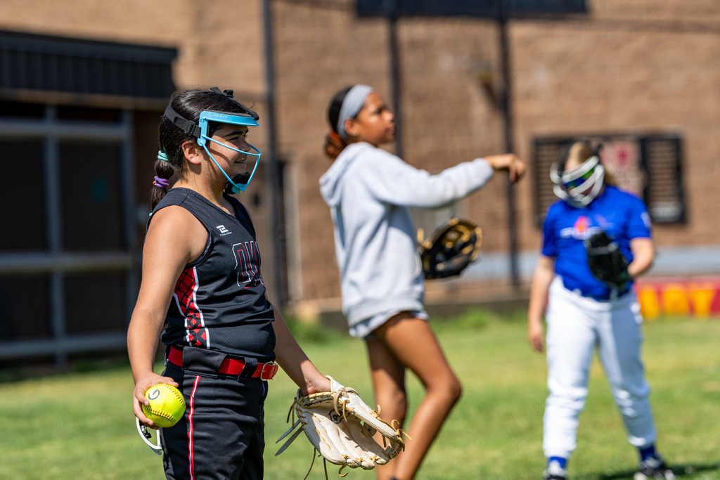 A student wearing a protective face mask holds a softball and glove as teammates warm up behind her.