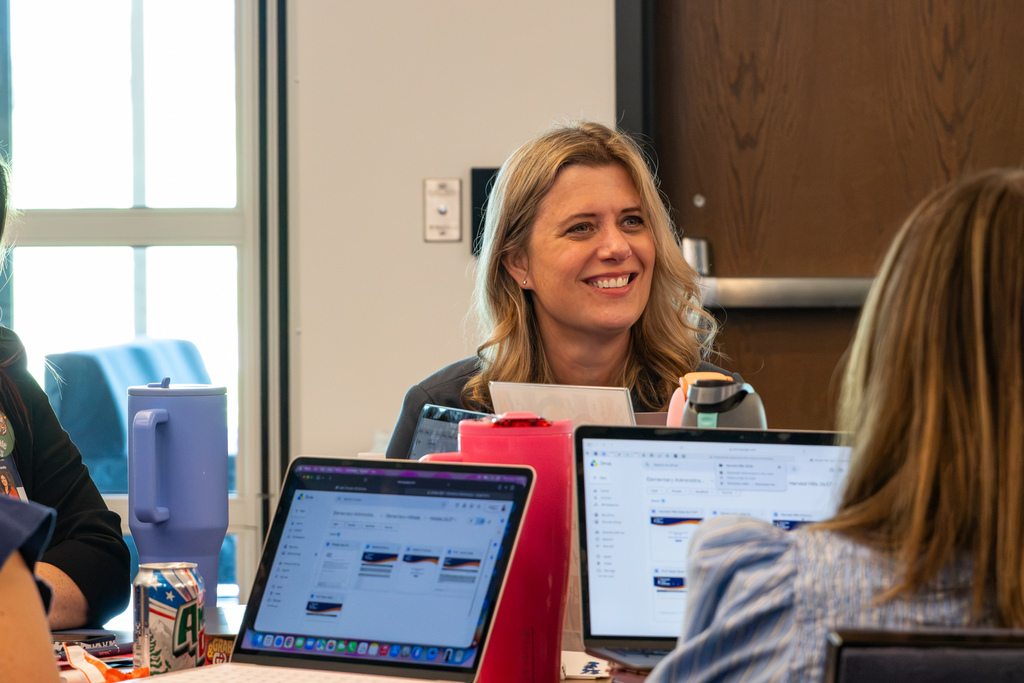 An administrator smiles while talking with colleagues seated around a table with laptops and workshop materials.