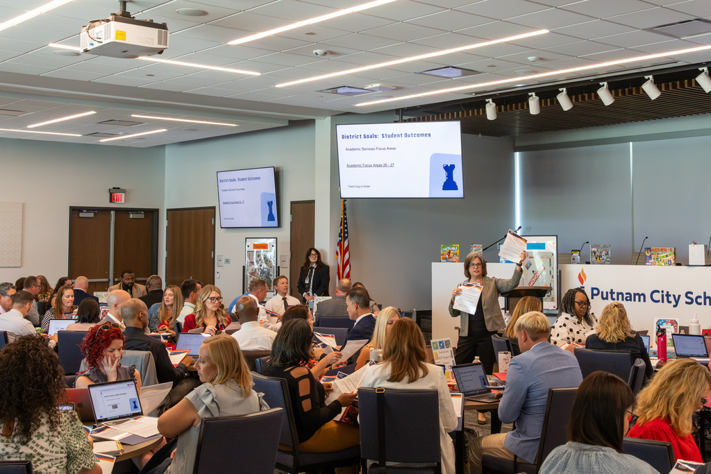 Administrators work in groups as a presenter holds up workshop materials at the front of the Centennial Center.