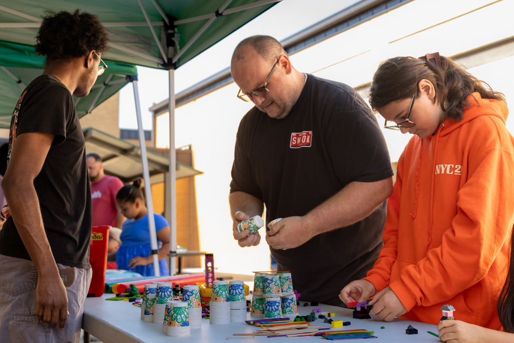 Students and adults build small creations with colorful bricks and craft supplies at an outdoor activity table.
