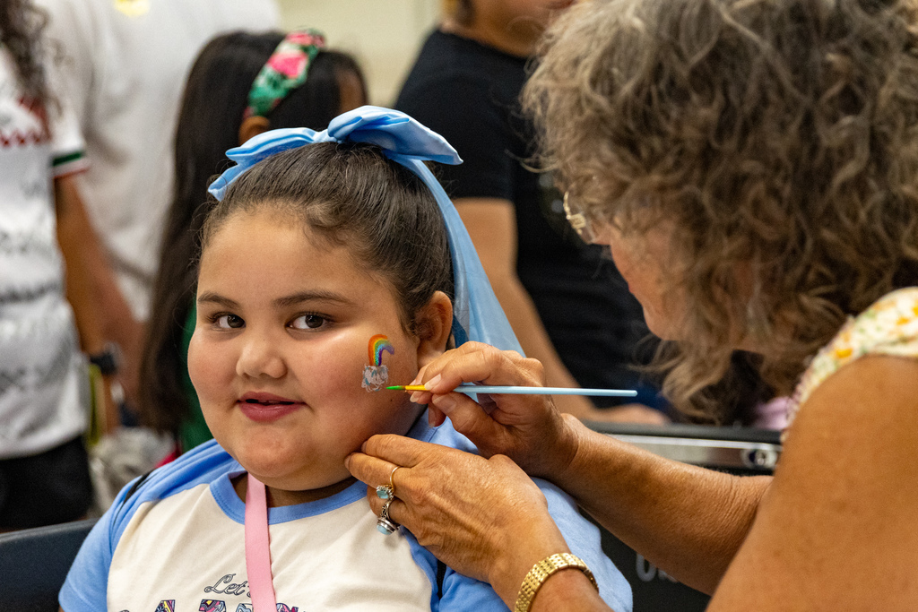 A child smiles while a volunteer paints a rainbow and clouds on her cheek.