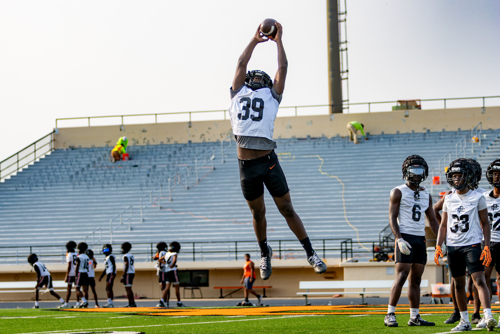 A Putnam City High football player leaps high and catches a pass above his head while teammates watch.