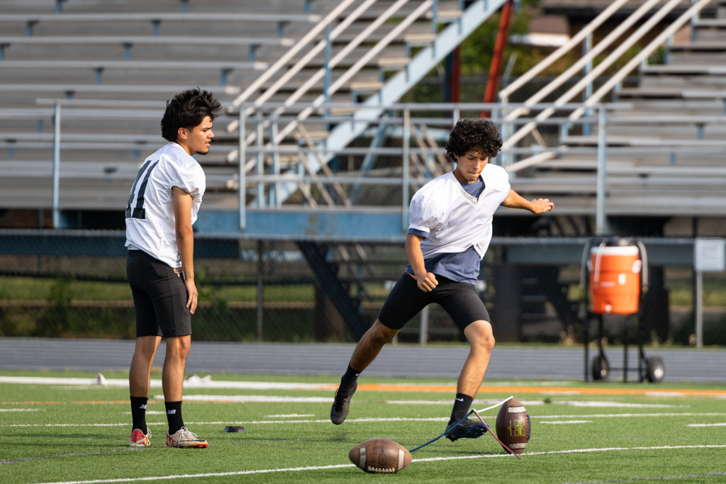 A Putnam City High player practices kicking a football from a tee while a teammate watches.
