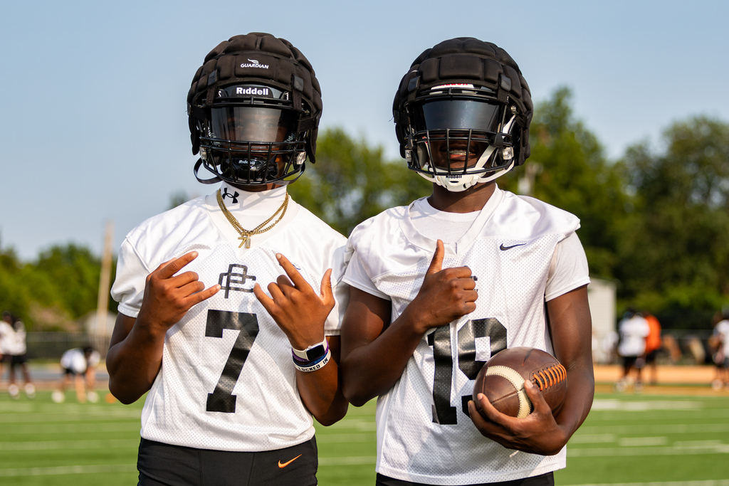 Two Putnam City High football players pose together in helmets and white practice jerseys, with one holding a football.