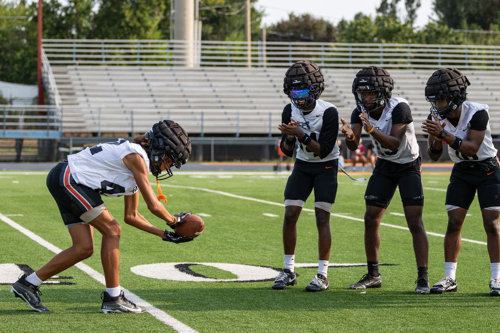 Putnam City High football players practice a snap drill as three teammates clap and wait in position on the field.