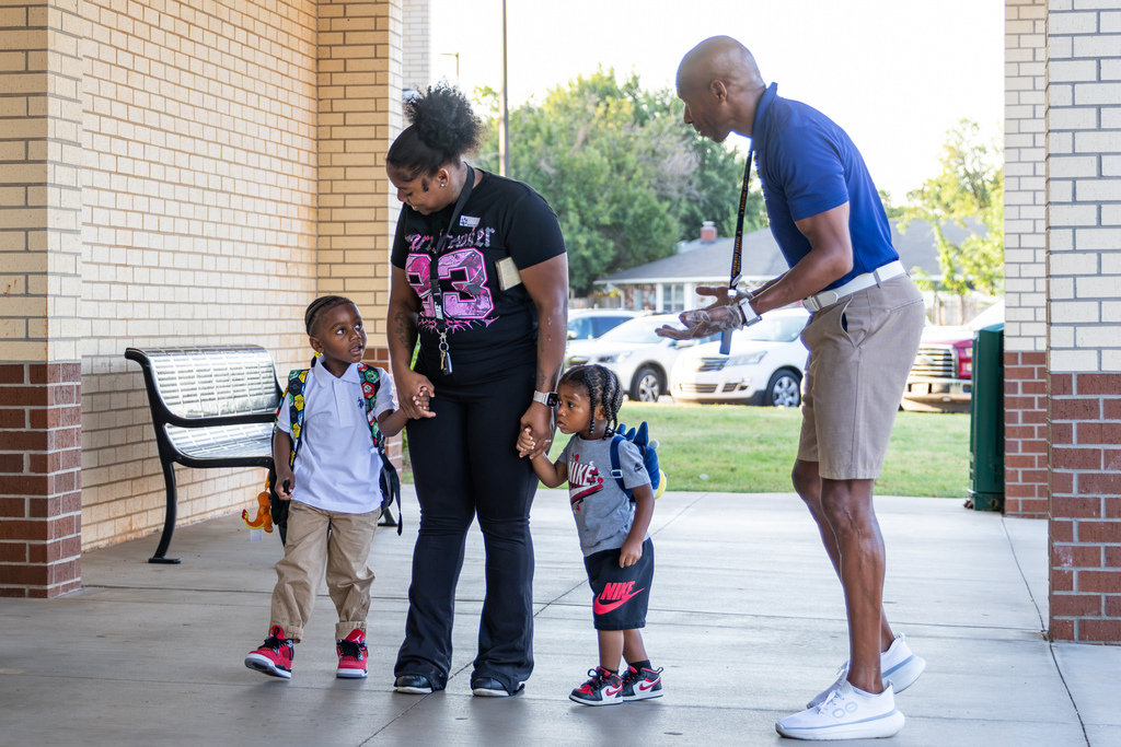 A school employee greets a family arriving for the first day as two young students stand beside their parent near the school entrance.