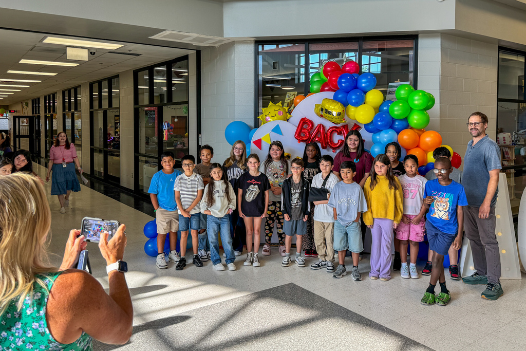 A large group of elementary students and a teacher pose together in front of a colorful “Back to School” balloon display inside the school.