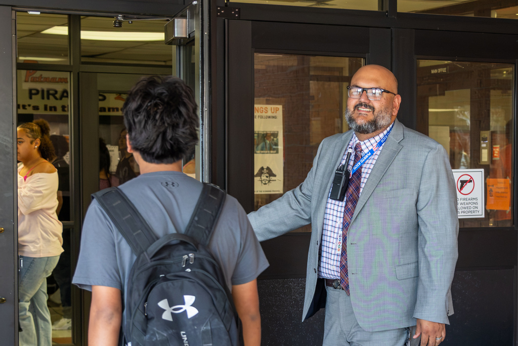 A school administrator smiles and holds the door open as a high school student with a backpack enters the building.