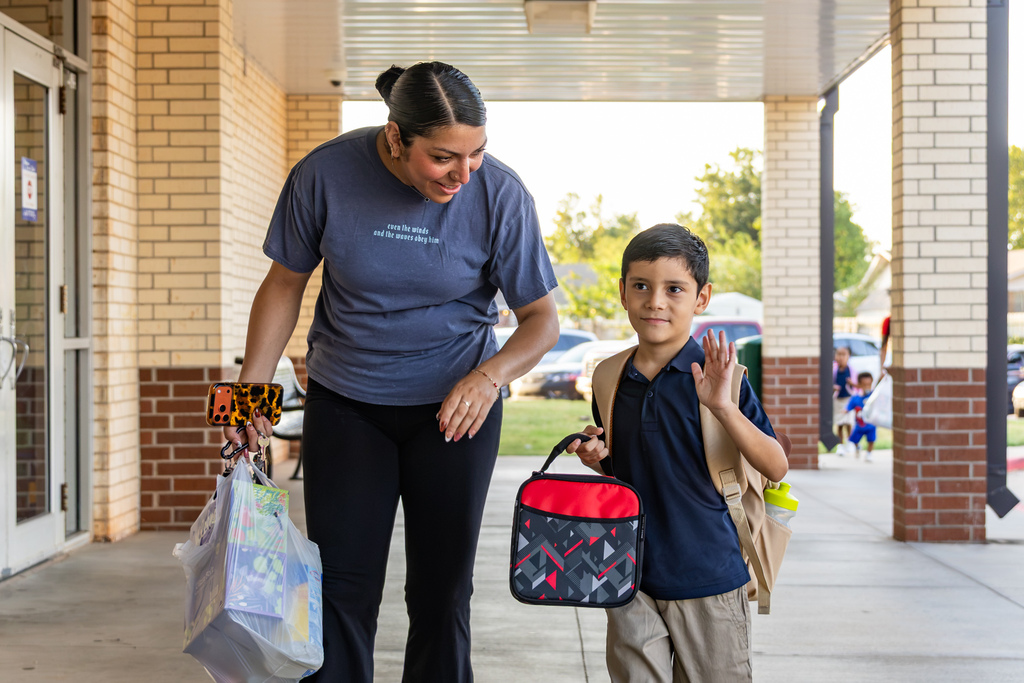 An elementary student carrying a lunchbox and backpack waves while walking into school beside an adult.