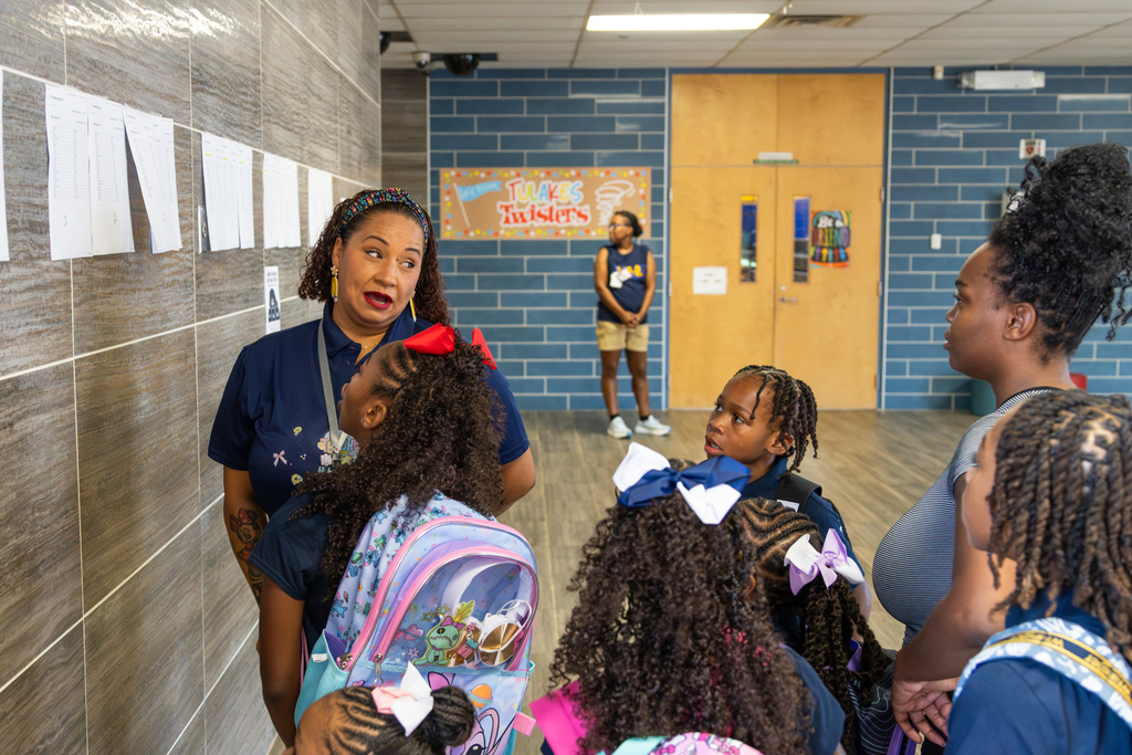 A school employee talks with several elementary students and a parent inside the school near posted class lists.