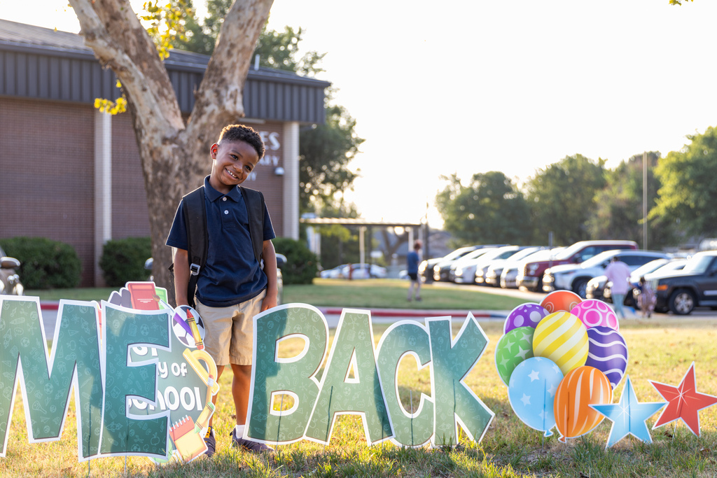 A smiling elementary student with a backpack stands behind a large colorful “Welcome Back” school display outside in the morning sunlight.