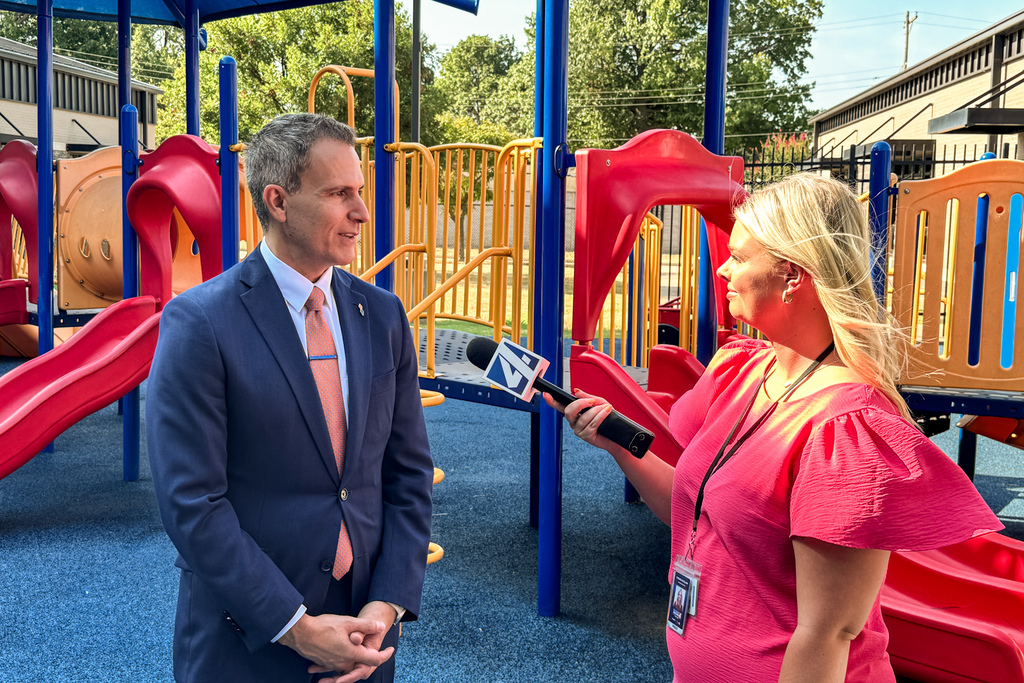 Superintendent Dr. Kenny Rodrequez speaks with a television reporter holding a microphone on a colorful school playground.