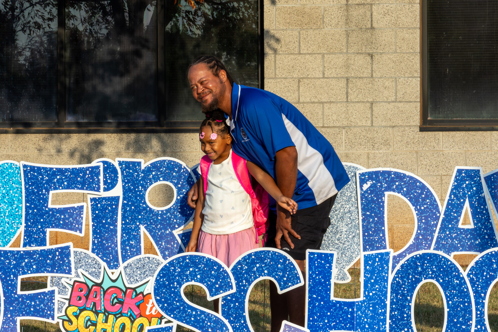 A father leans in beside his smiling young daughter as they pose together in front of a large blue “First Day of School” display.