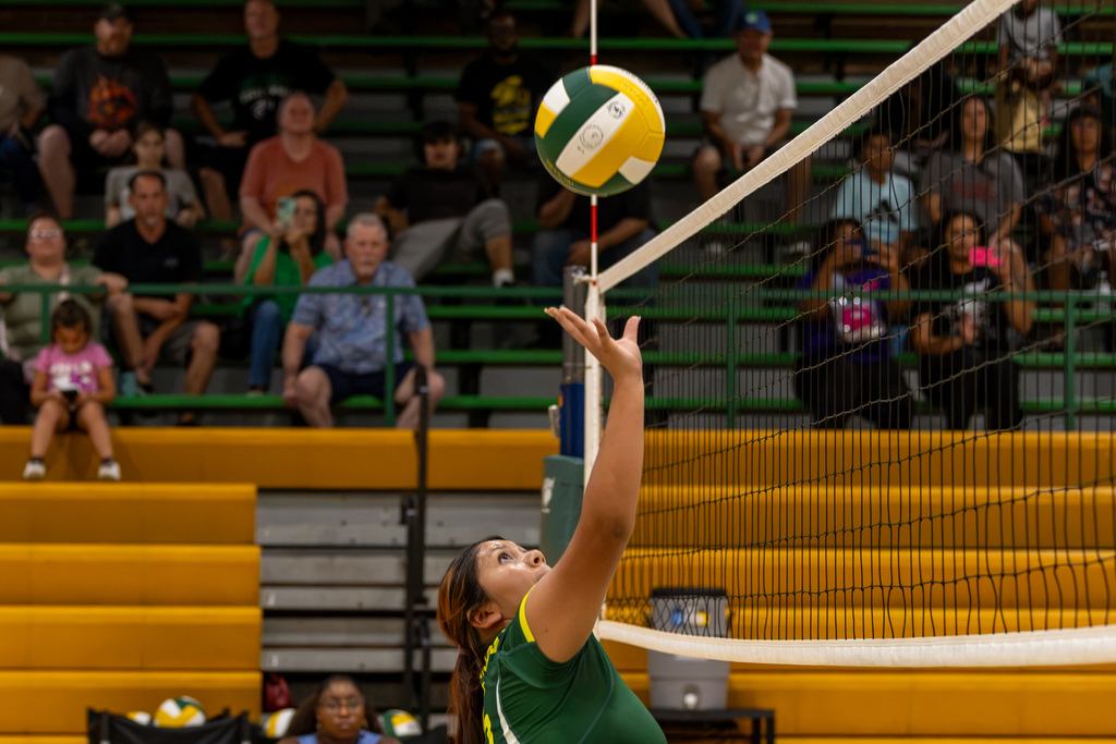 A Western Oaks Middle School volleyball player reaches up near the net as the ball drops above her hand, with spectators seated in the bleachers behind her.