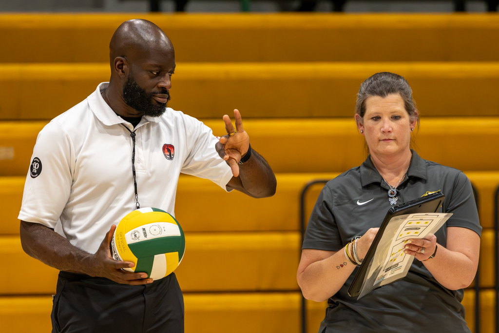 A Cooper coach stands courtside during the match near the referee, one holding a volleyball while the other reviews a clipboard.