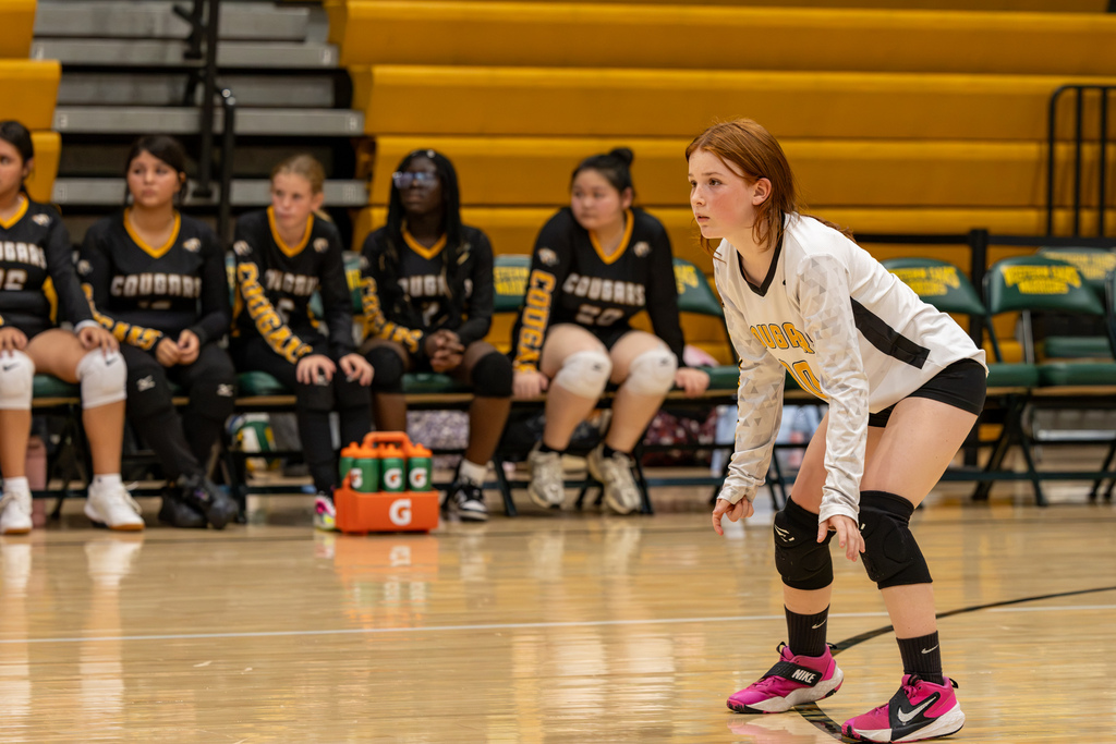 A Cooper Middle School volleyball player in a white libero jersey crouches in a ready position while teammates sit on the bench behind her.