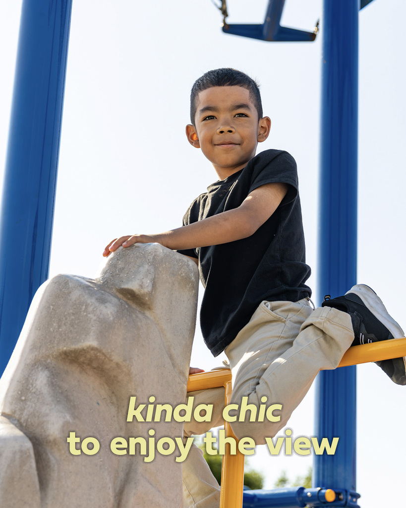 A smiling student sits high on a piece of playground equipment and looks out over the playground. Text reads, “kinda chic to enjoy the view.”