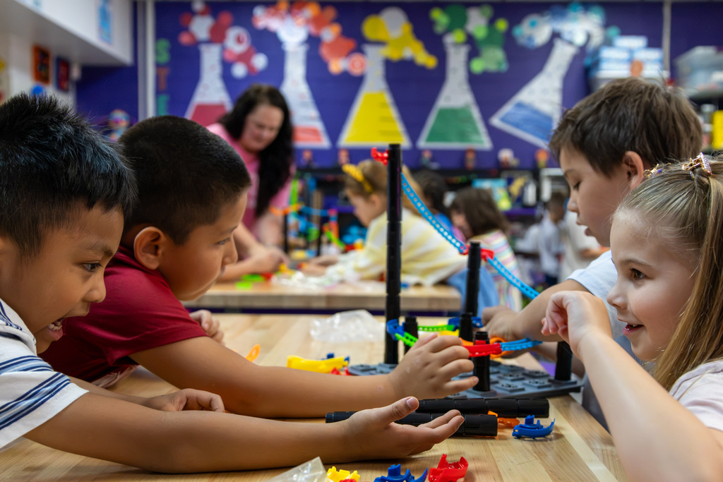 A group of Rollingwood Elementary students leans across a table while building a colorful roller coaster track, with more students working in the background.
