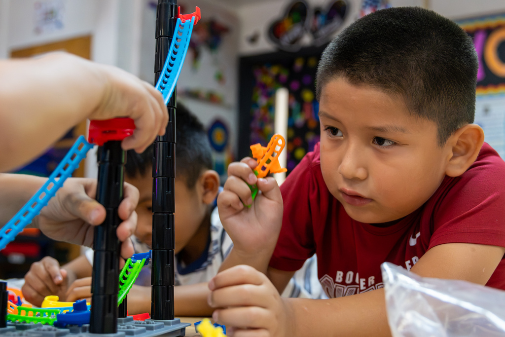 A Rollingwood Elementary student in a red shirt studies a roller coaster track up close while classmates add pieces nearby.