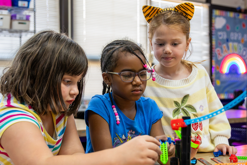 Three Rollingwood Elementary students concentrate on assembling roller coaster track pieces around a tall blue section of track.