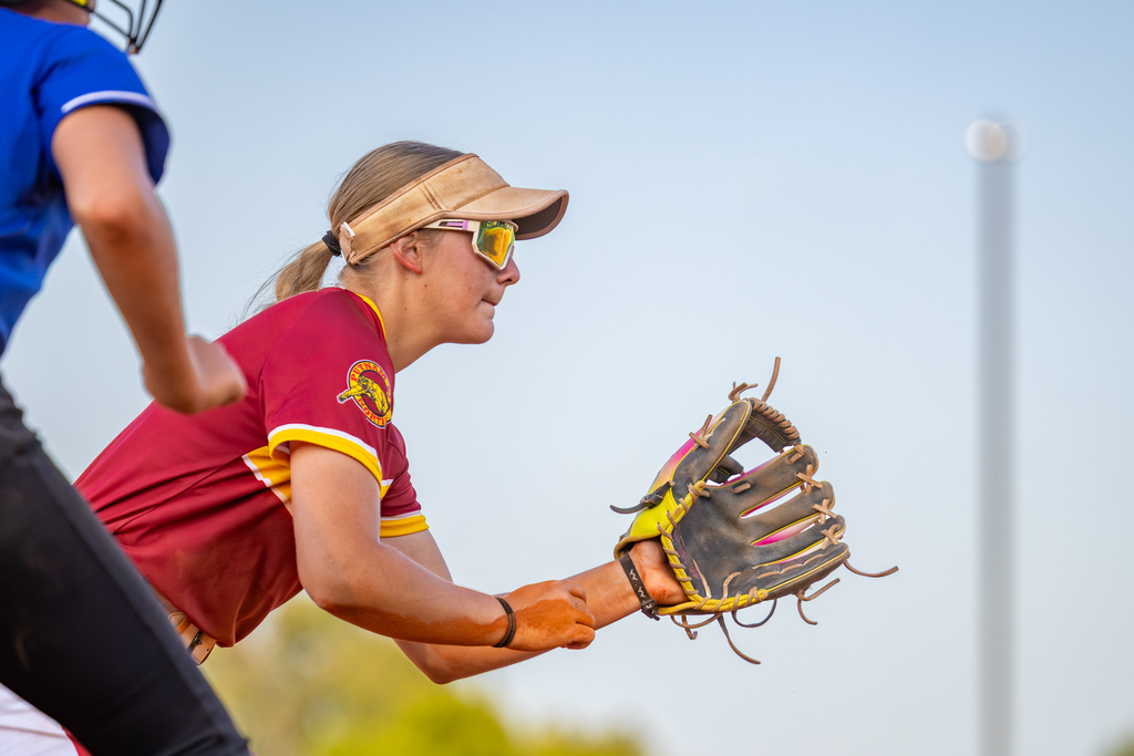 A PC North softball player crouches in the field with her glove open and ready as an opposing runner leads off in the foreground.