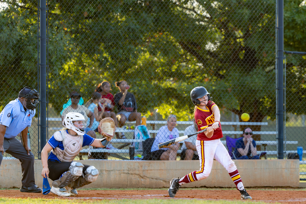 A PC North batter swings at a pitch as the opposing catcher reaches her glove toward the ball and the umpire watches from behind the plate.