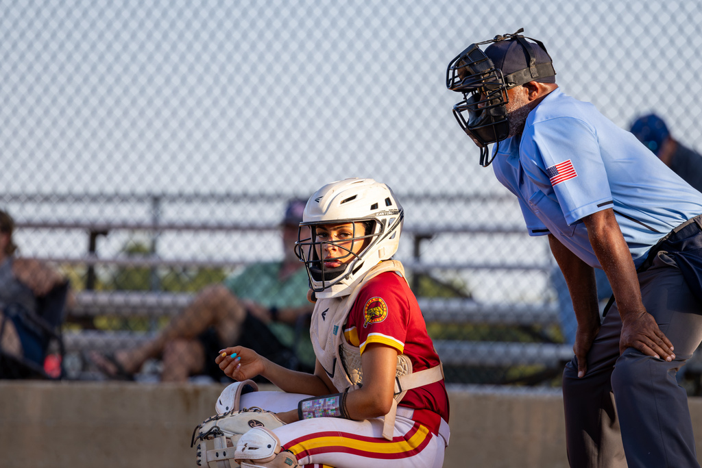 A PC North catcher kneels behind home plate and looks toward the field while the home plate umpire stands beside her.