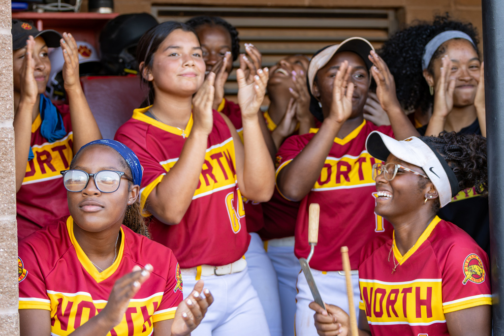 PC North softball players clap and cheer together from the dugout, with one player holding a drumstick as the team keeps the energy up.