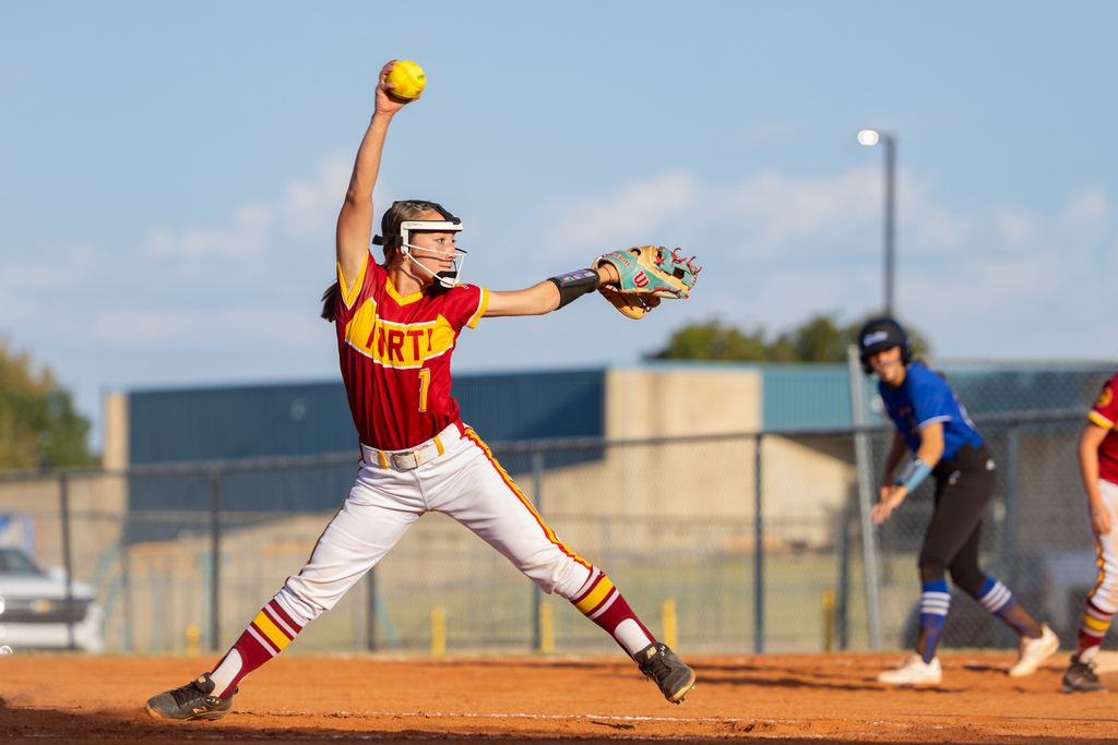 A PC North pitcher strides forward with her throwing arm raised high as she delivers a pitch toward home plate.