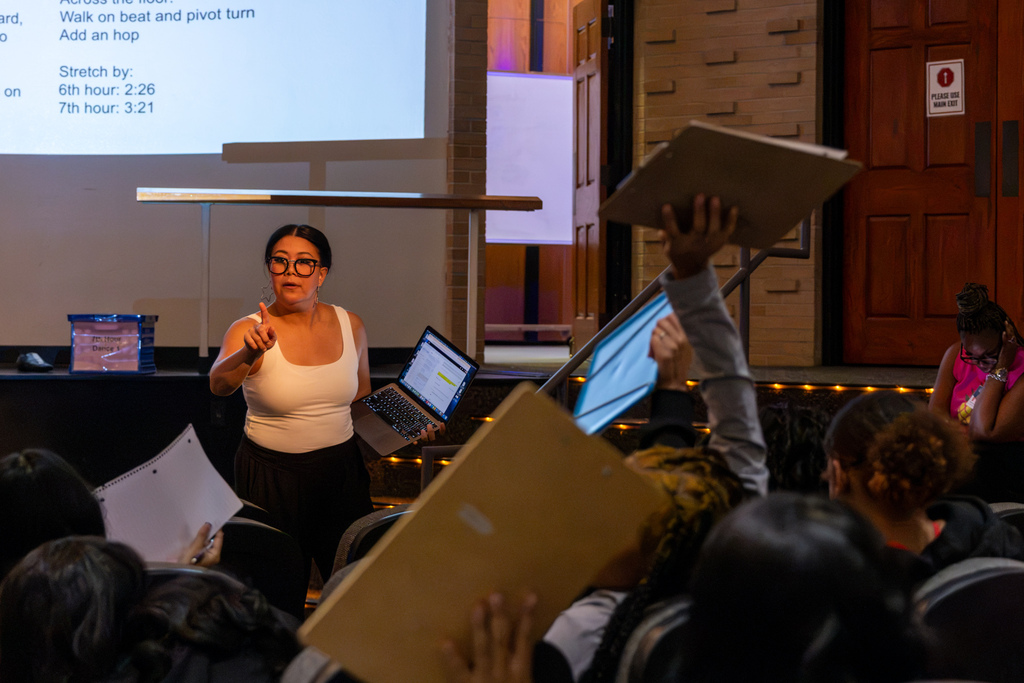 A PC High dance teacher speaks to students seated in the auditorium as several students raise clipboards during a class discussion.