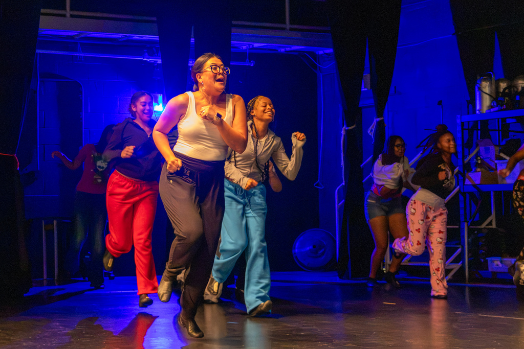 A PC High dance teacher and students smile while moving together under blue stage lighting during dance practice.