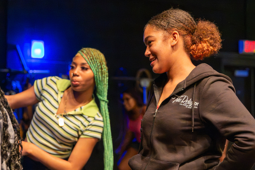 Two PC High dance students smile and talk together backstage during class, with blue stage lighting in the background.