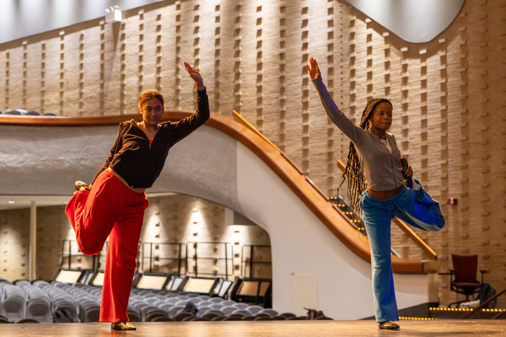Two PC High dance students balance on one leg with one arm raised while practicing movement on the auditorium stage.
