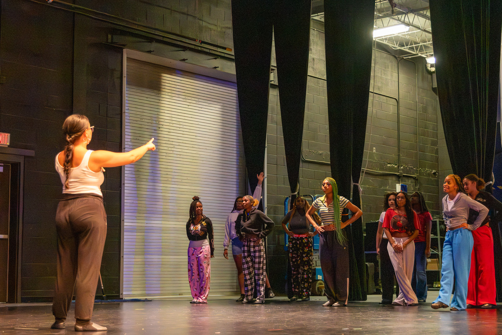 A PC High dance teacher gives instructions from the stage as students stand together backstage and listen.