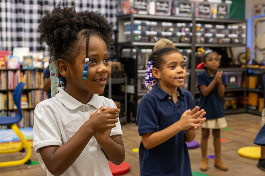 A first-grade student watches attentively with her hands clasped while classmates participate in a music activity behind her.