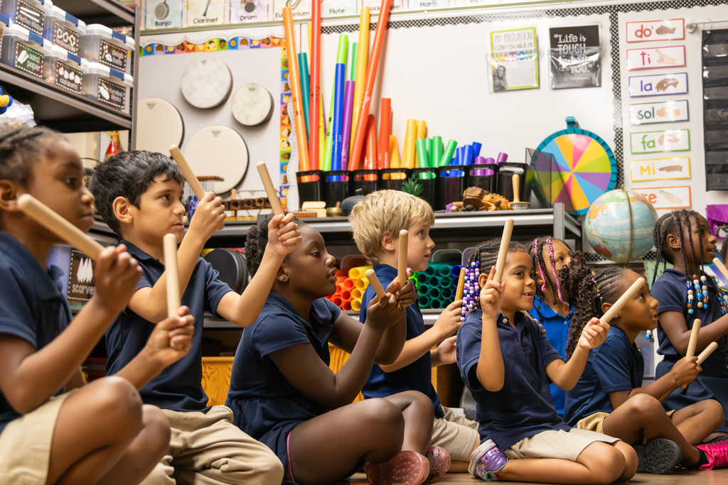 First-grade students sit on the music room floor holding rhythm sticks, with one student in the foreground preparing to tap hers together.