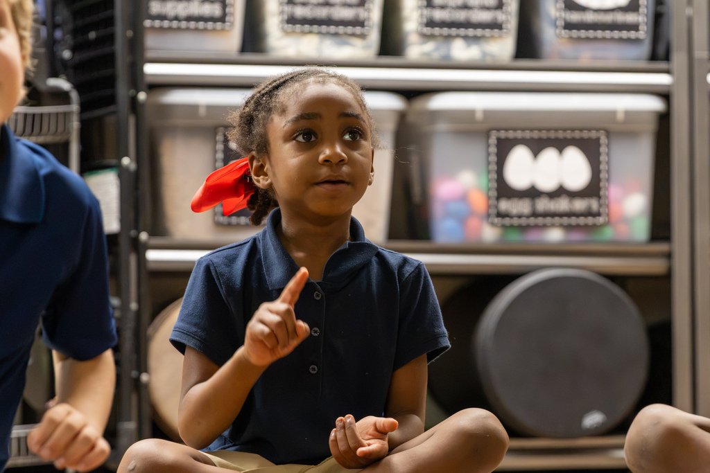 A first-grade student wearing a red hair bow sits on the floor and raises one finger while looking toward her teacher.
