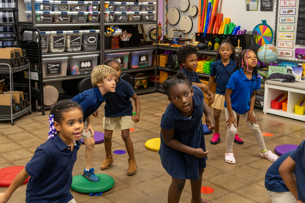 First-grade students move around colored floor spots during a music activity in a classroom filled with instruments and supplies.