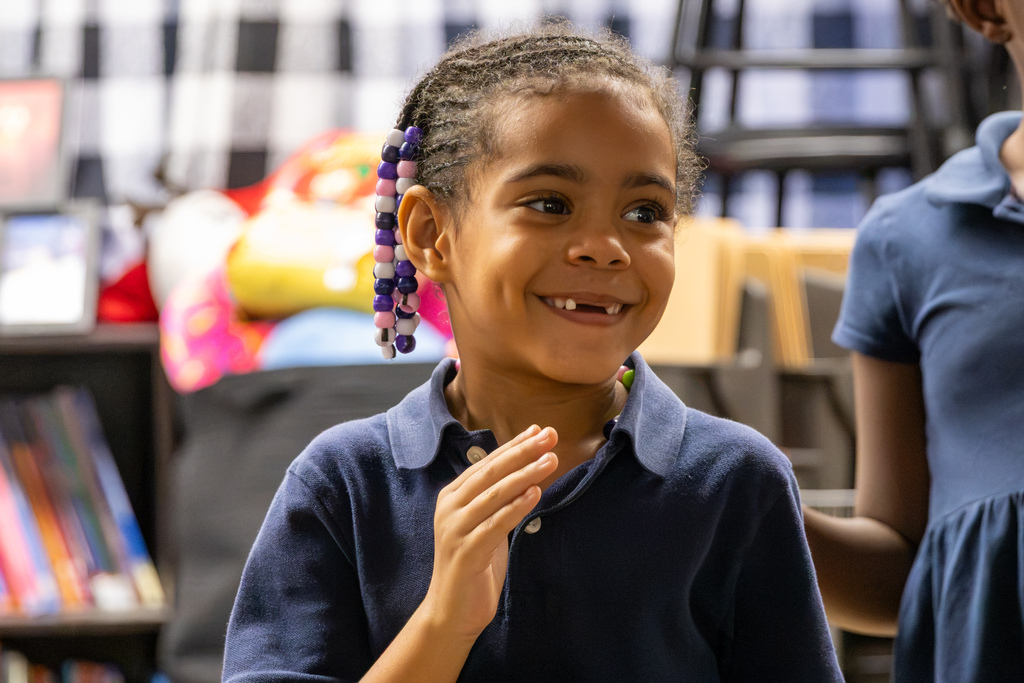 A smiling first-grade student with beaded braids participates in music class while another student is partially visible beside her.