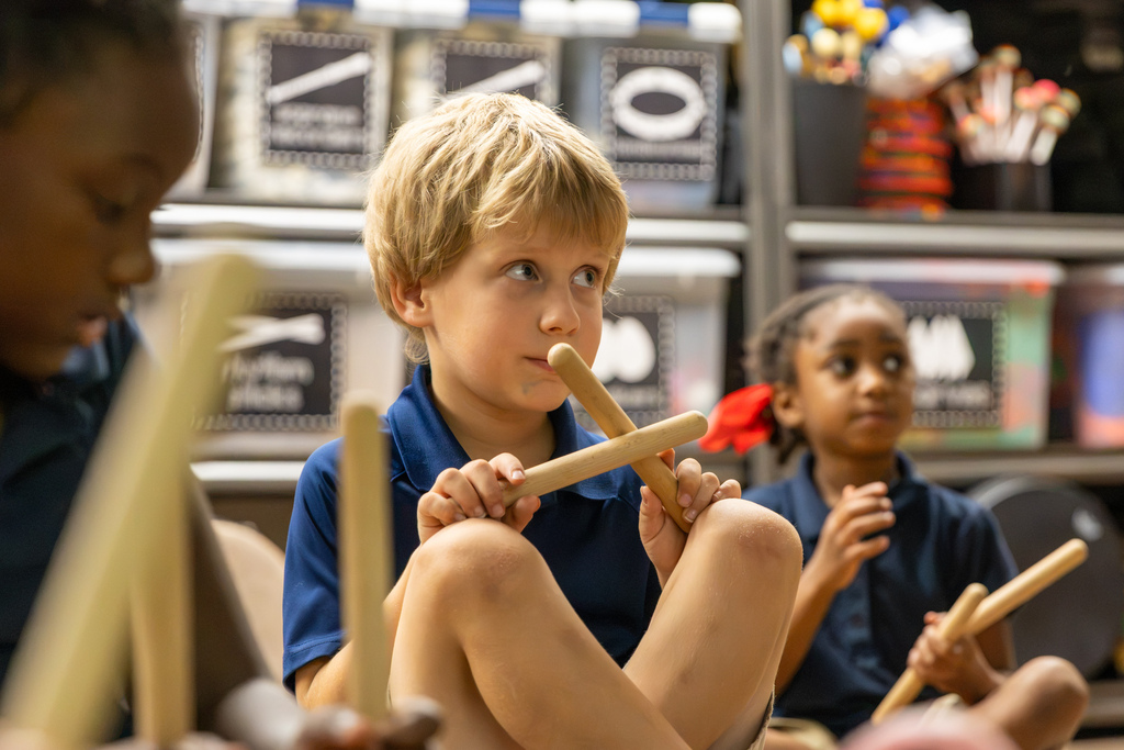 A first-grade student sits cross-legged and holds two wooden rhythm sticks together while classmates participate behind him.
